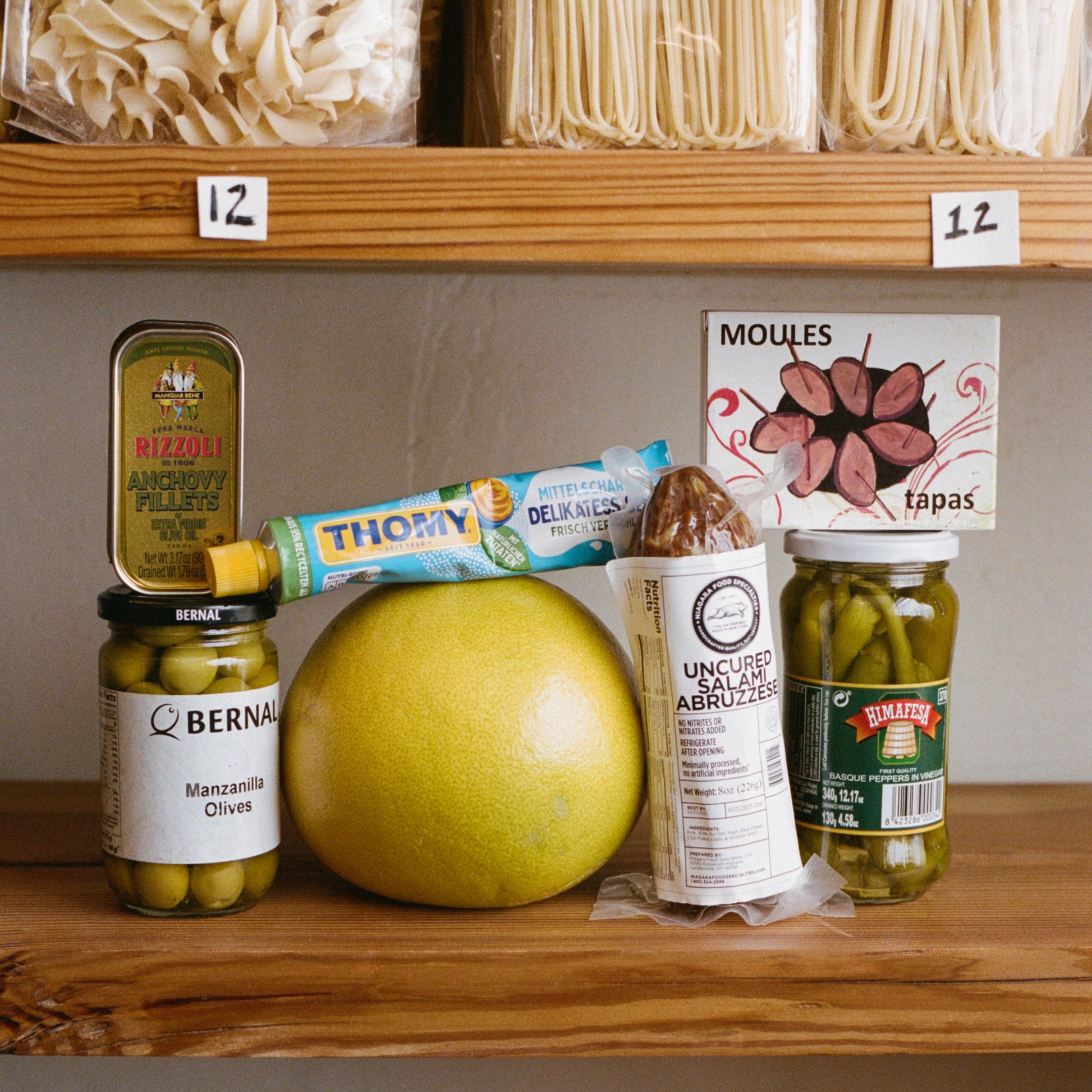 Assorted food items including a green fruit, jars, and packages on a wooden shelf.
