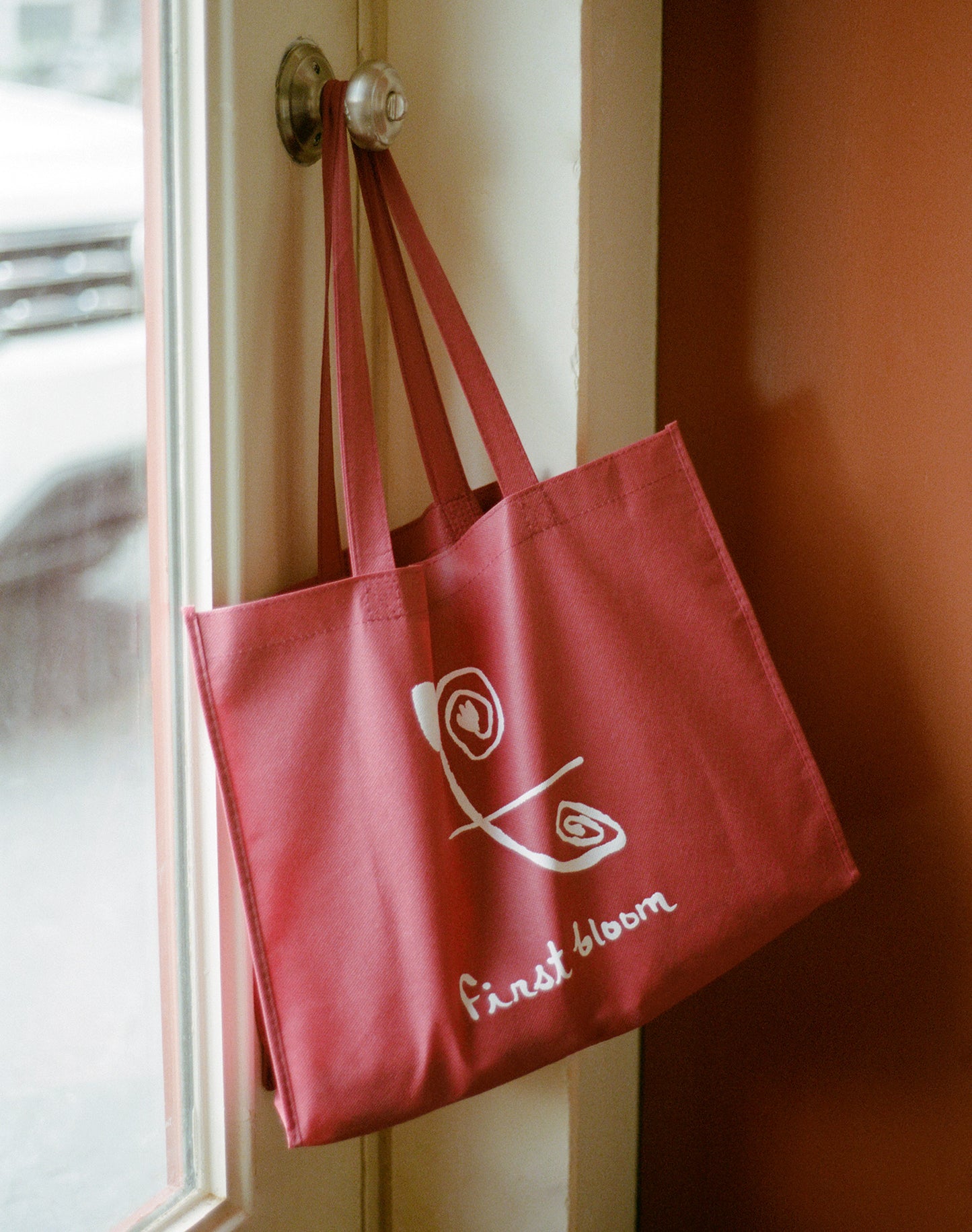 A red reusable shopping tote with white 'First Bloom' logo printed on it, hanging by a door knob.