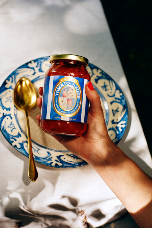 Hand holding a jar of tomato sauce with a decorative label on a table with a blue and white plate and gold spoon.