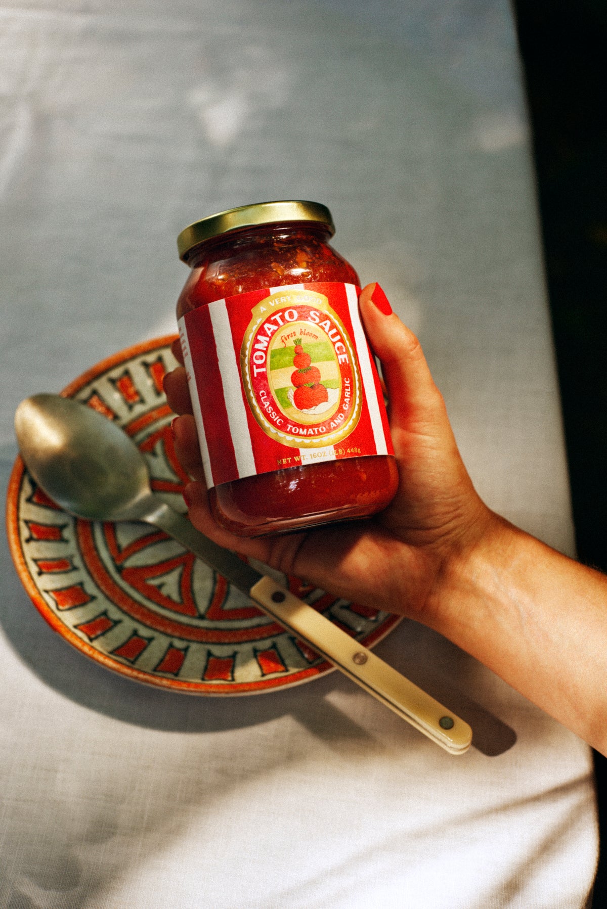 Hand holding a jar of tomato sauce with a spoon and decorative plate in the background