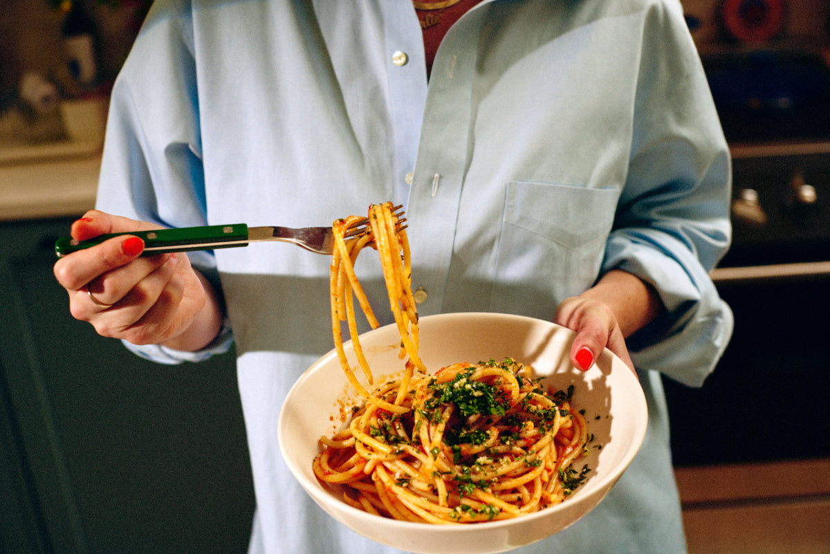 Person holding a bowl of spaghetti with a fork in a kitchen setting