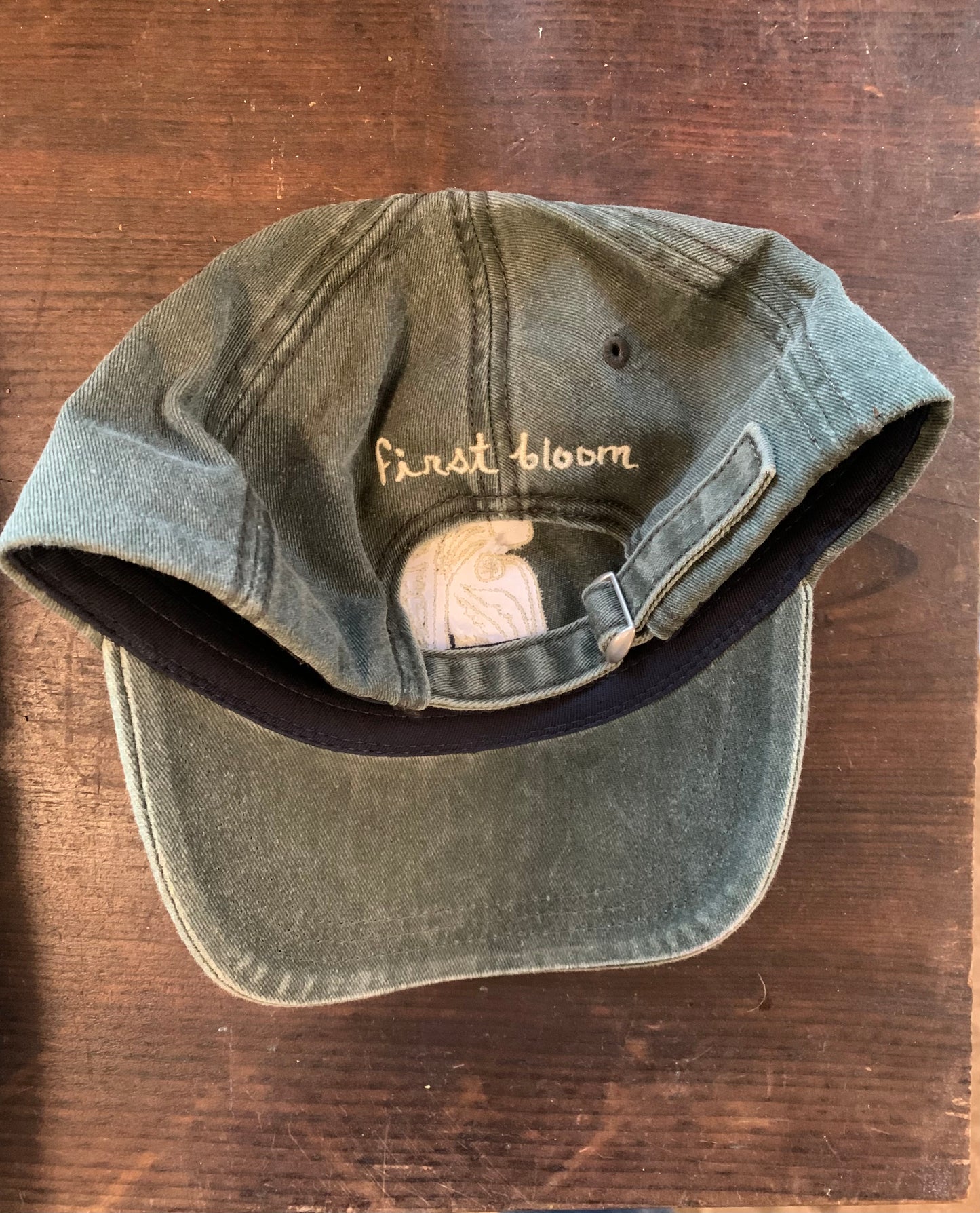 Back view shot of green baseball hat with first bloom logo text on the back, displayed on a wooden surface.