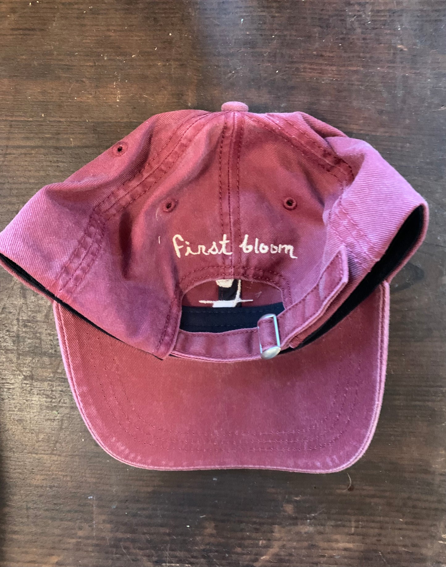 Back shot of a maroon baseball hat with white text and a logo on the front, displayed on a wooden surface.
