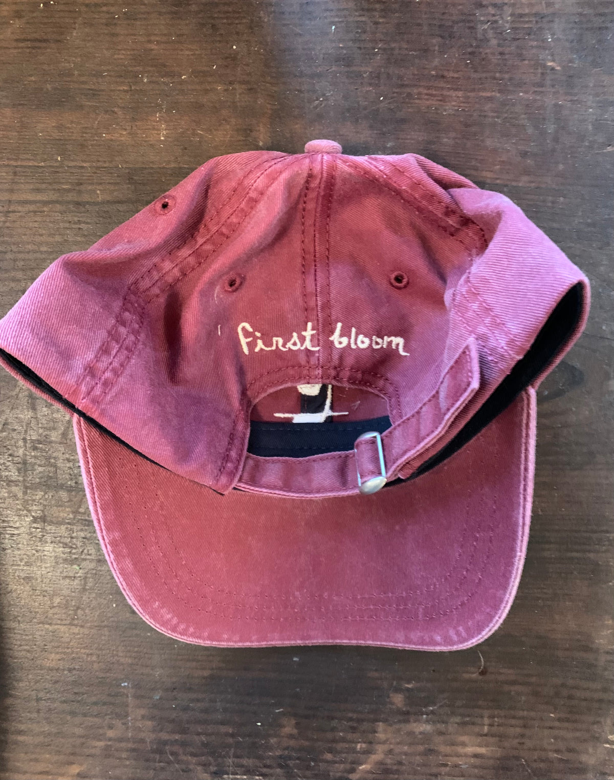 Back shot of a maroon baseball hat with white text and a logo on the front, displayed on a wooden surface.