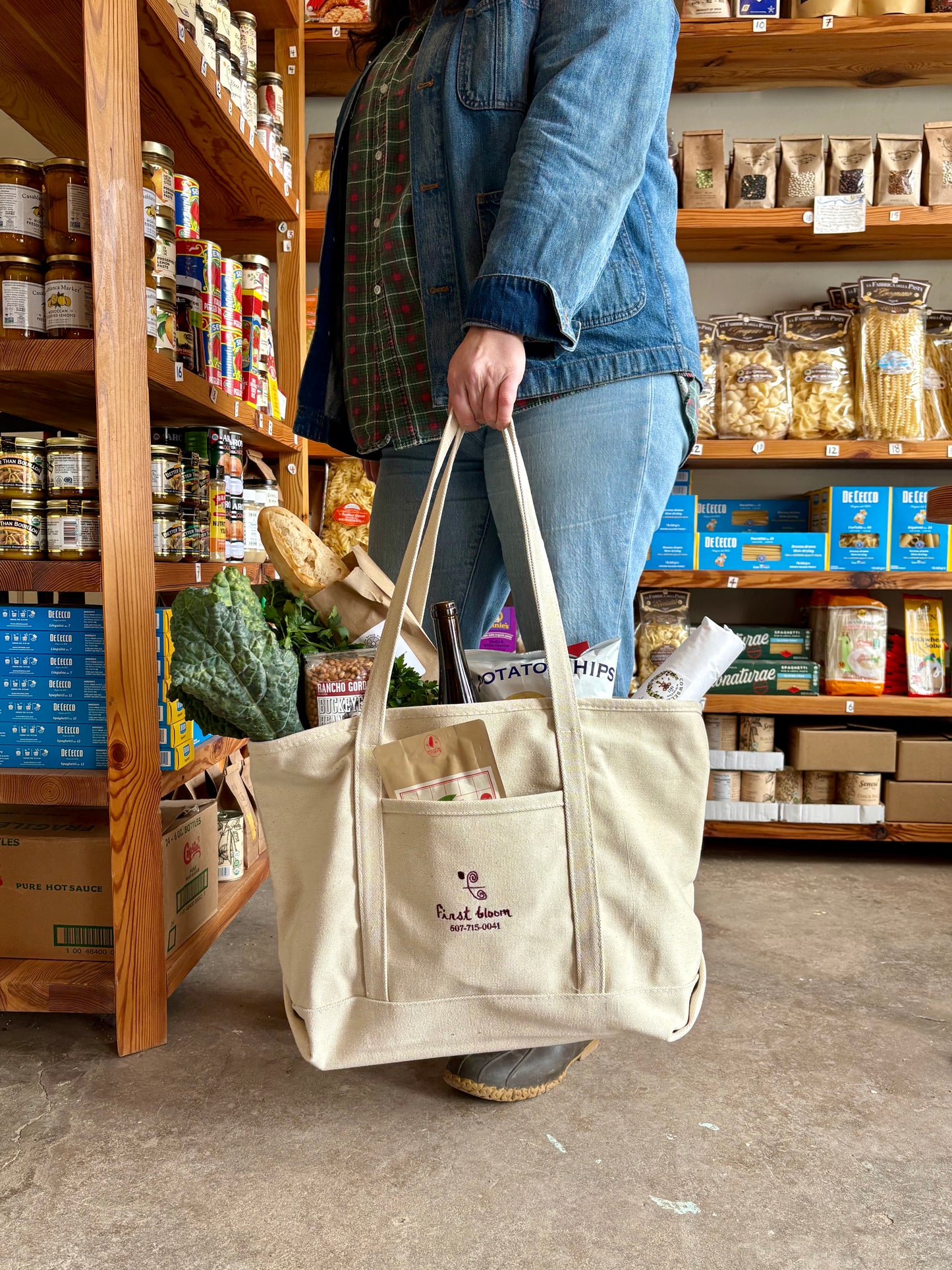 A person holding a beige canvas market tote with two handles and a front slip pocket, standing in a store.